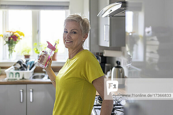 Lächelnde Frau hält Wasserflasche in der Küche zu Hause