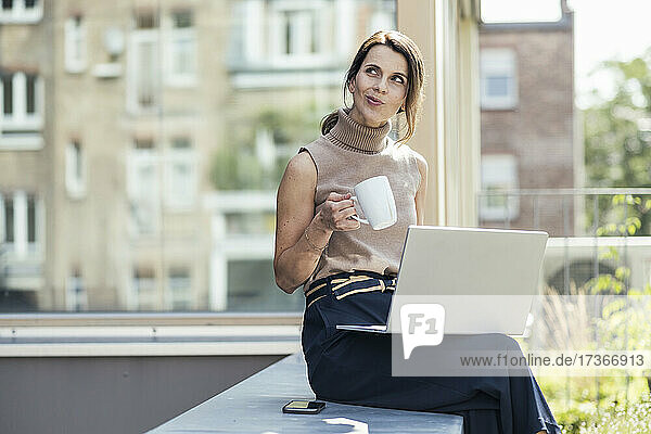 Nachdenkliche Geschäftsfrau sitzt mit Laptop und Kaffeetasse auf einer Stützmauer