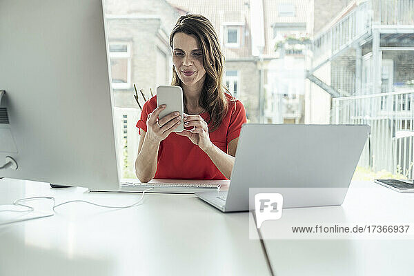 Weiblicher Berufstätiger  der am Schreibtisch sitzend ein Mobiltelefon benutzt