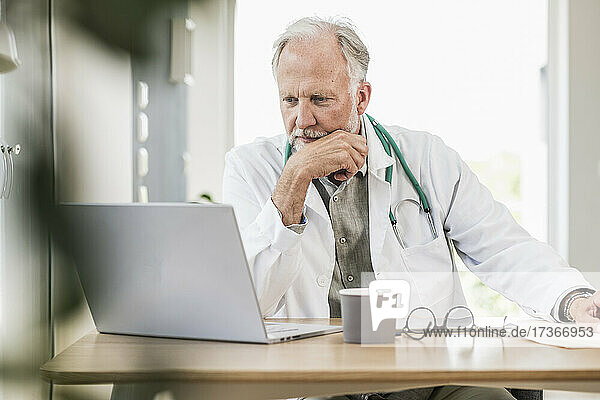 Reifer männlicher Arzt  der auf einen Laptop am Schreibtisch schaut