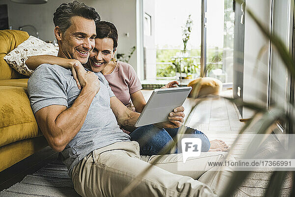 Lächelnde Frau mit einem Arm um einen Mann  der auf ein digitales Tablet schaut  während er zu Hause sitzt
