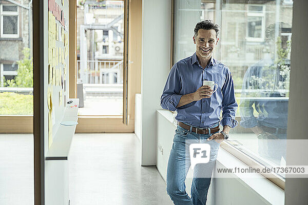 Lächelnder Geschäftsmann  der eine Kaffeetasse hält  während er im Büro steht