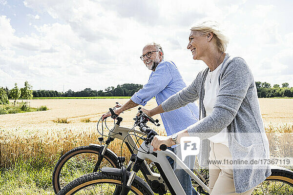 Smiling couple with bicycles walking by field during sunny day