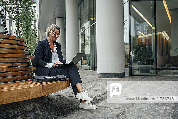 Lächelnde Geschäftsfrau  die einen Laptop benutzt und auf einer Bank im Büropark sitzt