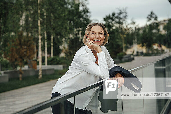 Lächelnde reife Geschäftsfrau mit Hand am Kinn  die sich auf ein Geländer stützt