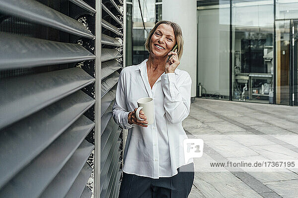 Geschäftsfrau mit Tasse  die mit ihrem Smartphone telefoniert  während sie an den Jalousien im Büropark steht