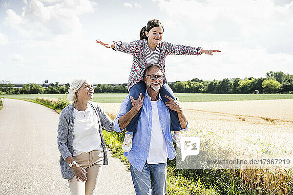 Grandfather carrying cheerful granddaughter on shoulder while walking by woman on road