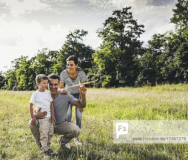 Father holding airplane toy while crouching by son and woman on grass