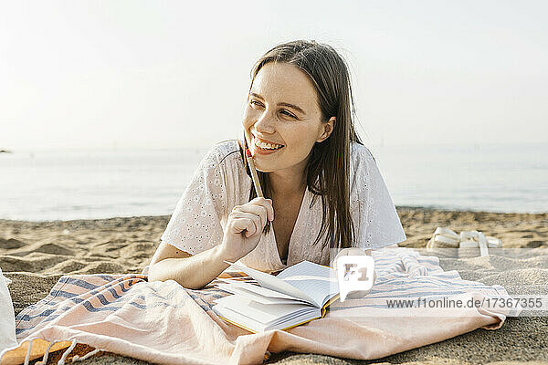 Nachdenkliche glückliche Frau  die einen Stift hält  während sie sich am Strand ausruht