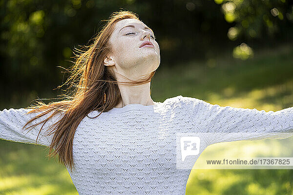 Junge Frau mit geschlossenen Augen streckt ihre Arme im Park aus