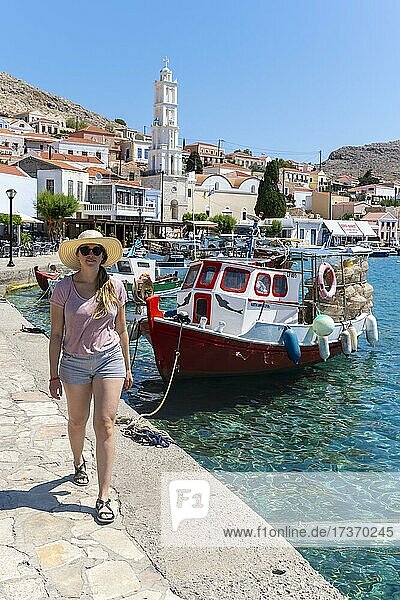 Touristin mit Sonnenhut  Fischerboote im Hafen von Chalki mit türkisblauem Wasser  Promenade mit bunten Häusern des Ortes Chalki  Chalki  Dodekanes  Griechenland  Europa