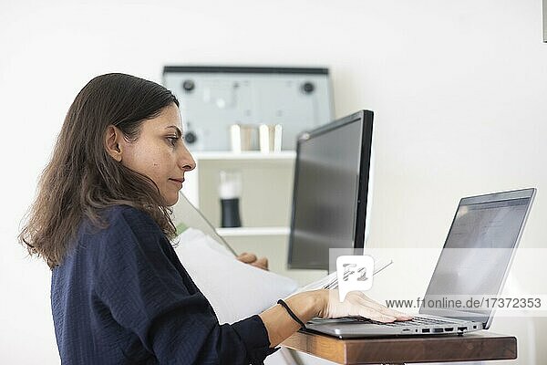 Mittelalte Frau mit dunklen Haaren kontrolliert Produkt bei der Arbeit mit Laptop und Ordner  Freiburg  Baden-Württemberg  Deutschland  Europa