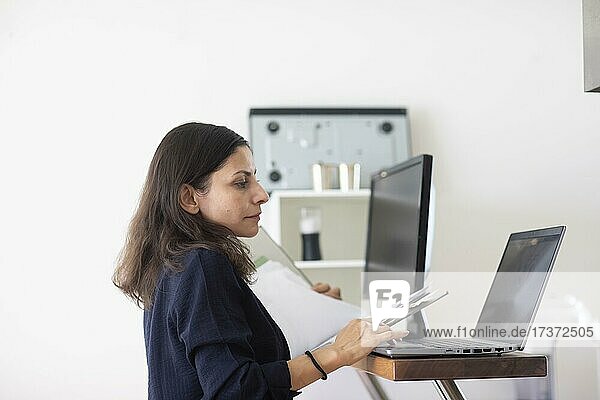 Mittelalte Frau mit dunklen Haaren kontrolliert Produkt bei der Arbeit mit Laptop und Ordner  Freiburg  Baden-Württemberg  Deutschland  Europa