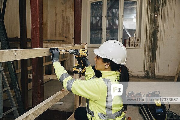 Weiblicher Arbeiter mit Schutzhelm bei der Arbeit auf einer Baustelle mit einer Bohrmaschine auf einem Holzbrett