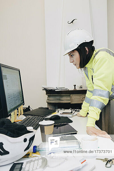 Weibliche Bauunternehmerin mit Schutzhelm verwendet einen Computer  während sie sich im Büro auf einen Schreibtisch stützt