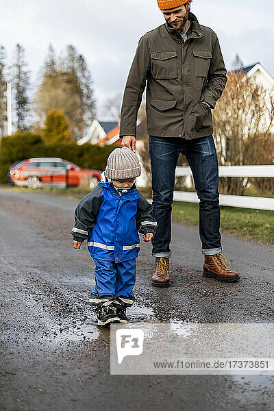 Full length of boy walking on puddle by father standing on road
