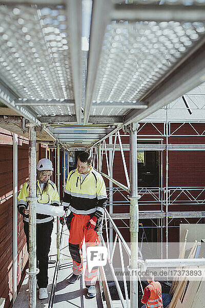 Bauunternehmerin in voller Länge  die mit einem männlichen Kollegen einen Grundriss bespricht  während sie auf einem Baugerüst läuft