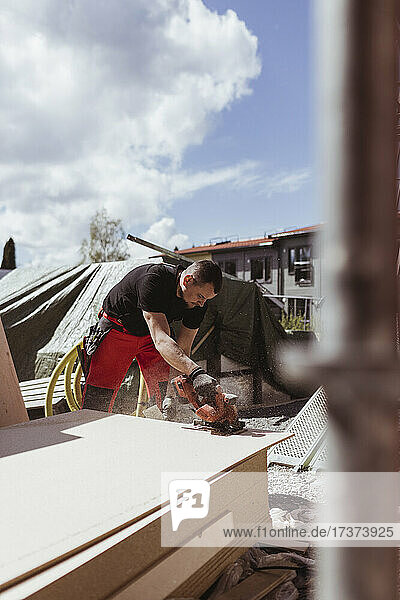 Männlicher Zimmermann  der an einem sonnigen Tag auf einer Baustelle mit einer elektrischen Säge Holzbretter schneidet