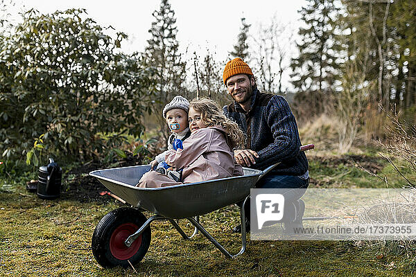 Boy and girl sitting in wheelbarrow by father at back yard