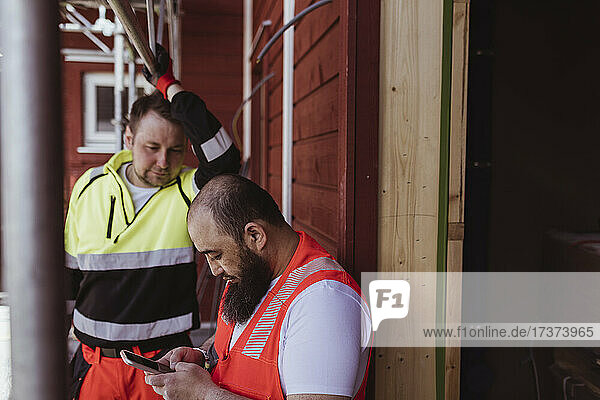 Männlicher Bauarbeiter schaut auf einen bärtigen Kollegen  der auf der Baustelle ein Smartphone benutzt
