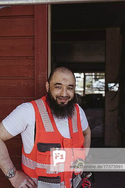Lächelnder bärtiger männlicher Bauarbeiter  der auf einer Baustelle steht und die Hand auf die Hüfte legt