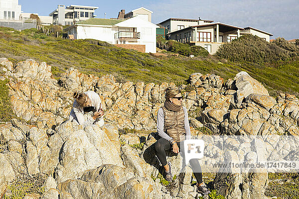 Mutter und Tochter im Teenageralter sitzen auf den Felsen unterhalb der Luxushäuser auf einer Landzunge