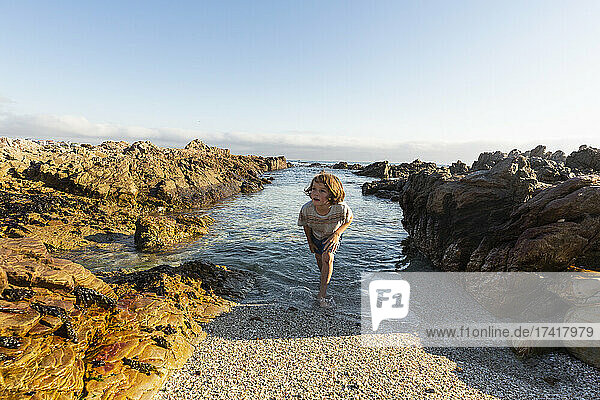 A young boy in shallow sea water among jagged rocks on the beach at De Kelders.