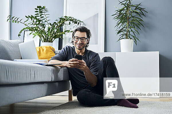 Smiling businessman using smart phone while leaning on sofa at home