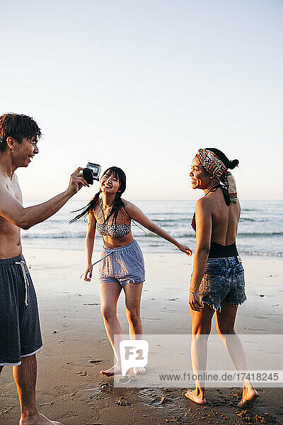 Fröhlicher junger Mann zeigt Freundinnen am Strand die Kamera
