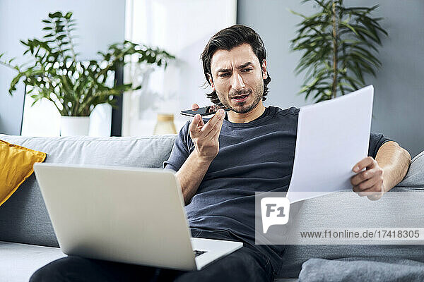 Businessman discussing on smart phone holding document while sitting with laptop at home