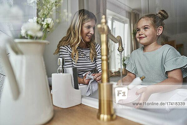 Smiling girl looking through window while washing dishes with mother in kitchen