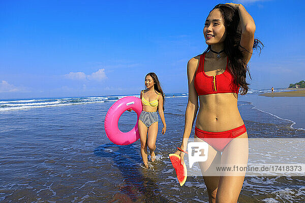 Smiling woman holding watermelon while female friend with swimming float walking through water on sunny day