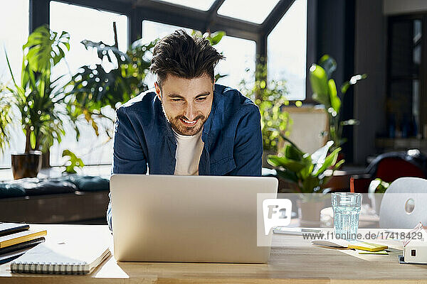 Smiling male professional using laptop while leaning on desk in office
