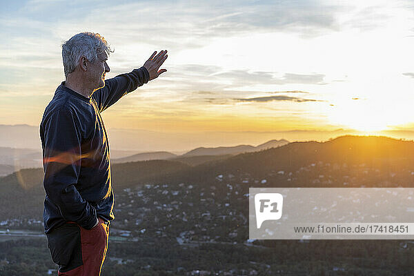 Senior man shielding eyes while standing on mountain during sunset