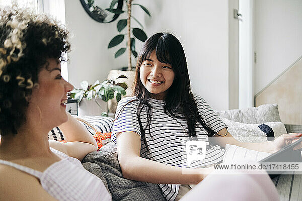 Happy woman with digital tablet looking at friend while sitting on sofa