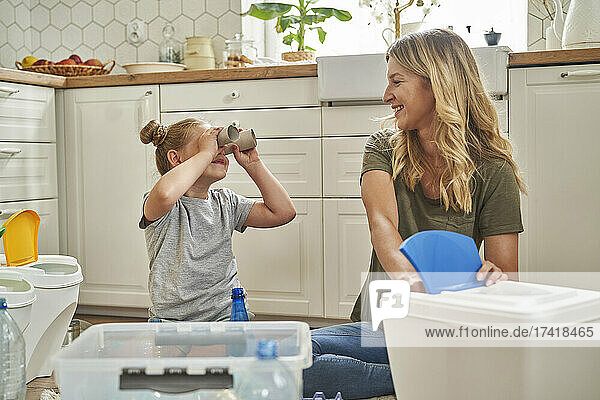 Playful girl looking at mother through rolled up paper in kitchen