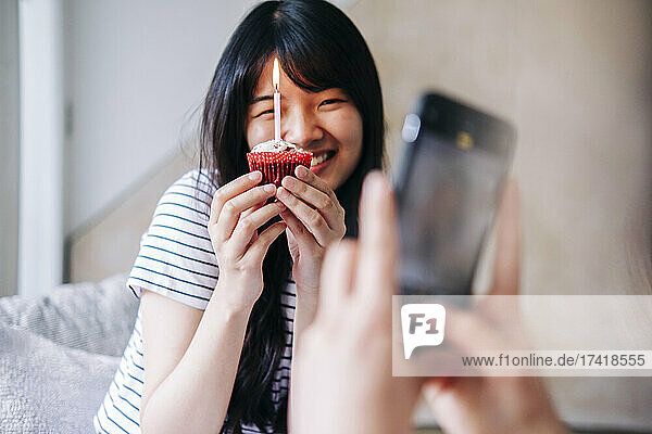 Woman taking photo of smiling friend with cupcake through mobile phone at home