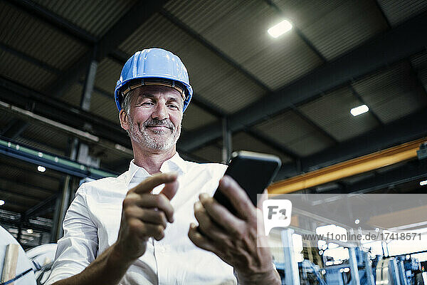 Smiling male engineer with hardhat using mobile phone at metal industry