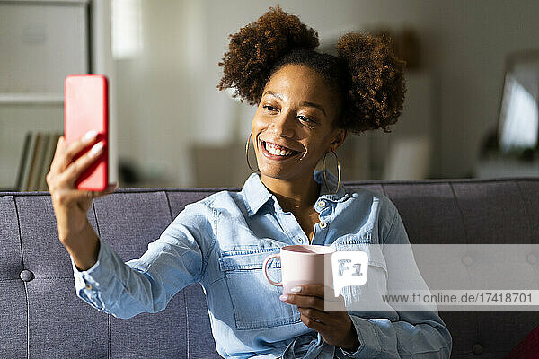 Smiling woman talking selfie with coffee cup through mobile phone at home
