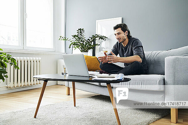 Männlicher Profi während eines Videoanrufs auf dem Laptop  während er zu Hause auf dem Sofa sitzt