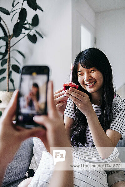 Woman photographing happy friend with cupcake through mobile phone at home