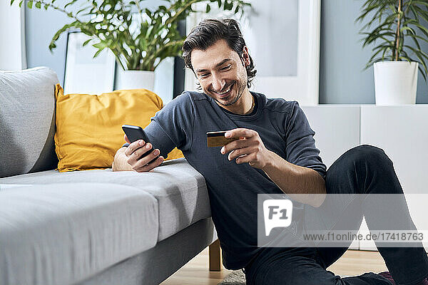 Smiling man with smart phone shopping online through credit card at home