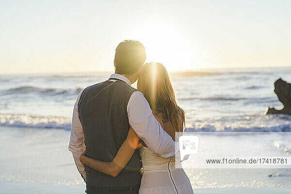 Young newlywed couple with arm around looking at sunrise view at beach