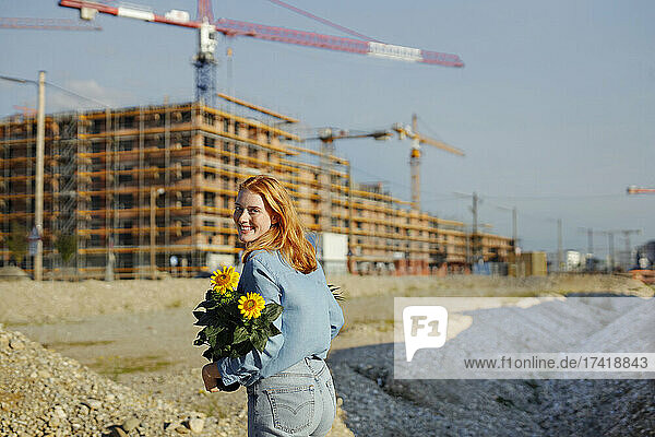Smiling woman with flowering plant walking at construction site on sunny day