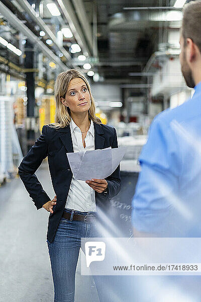 Female business professional with hand on hip looking at colleague in factory