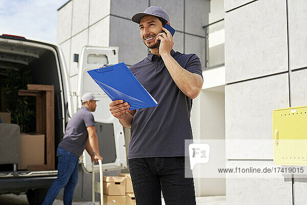 Smiling delivery man holding clipboard while talking on smart phone