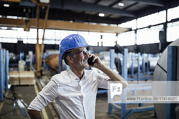 Smiling male engineer with hardhat talking on mobile phone in steel mill
