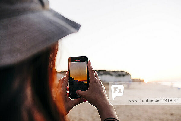 Woman photographing through smart phone at beach during sunset