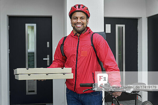 Lächelnder Lieferbote mit Rucksack hält Pizzakartons und Fahrrad vor dem Haus