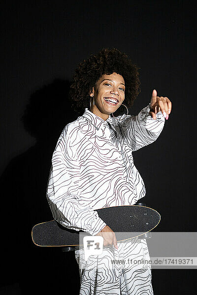 Female fashion model pointing while holding skateboard against black background
